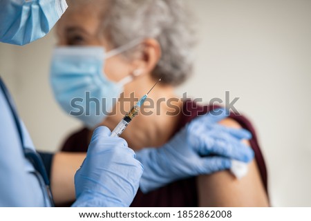 Close up of general practitioner hand holding vaccine injection while wearing face protective mask during covid-19 pandemic. Young woman nurse with surgical mask giving injection to senior woman.