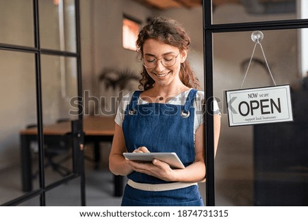 Happy waitress standing at restaurant entrance holding digital tablet to check the reservations. Smiling owner in blue apron standing at coffee shop entrance leaning on door with open signboard.
