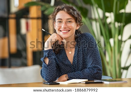 Portrait of smiling woman wearing spectacles while sitting at desk. Business woman taking notes in diary and looking at camera. University girl with eyeglasses sitting on table at library.