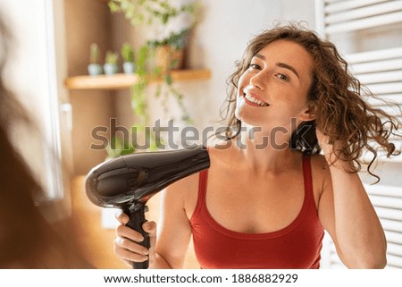 Beautiful girl using a hair dryer and smiling. Natural young woman drying curly hair with hair-dry machine. Happy beauty looking at mirror while using hair dryer in the bathroom after shower.