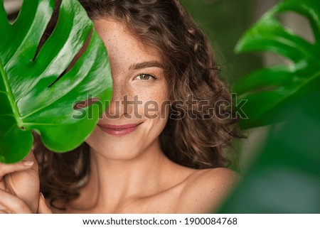 Close up face of beautiful young woman covering her face by green monstera leaf while looking at camera. Portrait of beauty woman with natural makeup and freckles standing behind big green leaves.