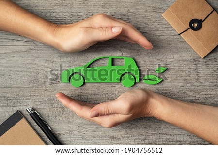 Close up of hands of woman protecting icon of green eco-friendly car over wooden table. Top view of woman hands protecting electric car with care, mobility. Car insurance and green automotive concept.