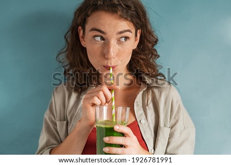 Beautiful woman drinking an organic green smoothie. Fit young woman drinking detox juice using paper straw isolated against blue background. Healthy girl enjoy detox drink and looking away. 