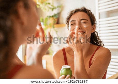 Playful young woman applying cream on nose. Girl holding green lotion jar applying moisturizer on nose. Beautiful woman taking care of skin by applying moisturizer every day in the morning.