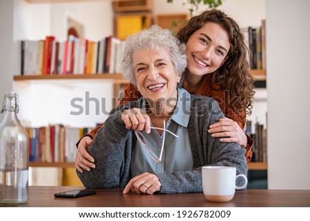 Young woman embracing senior mother at home and looking at camera. Portrait of happy adult granddaughter and grandmother embracing and smiling together. Lovely young woman hugging from behind grandma 