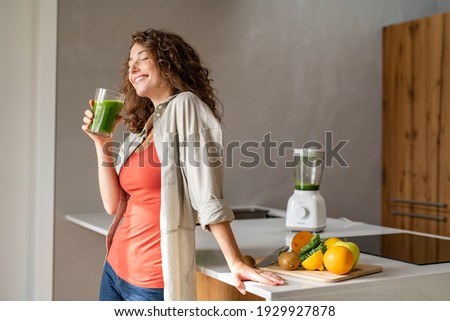 Happy young woman standing in kitchen and holding glass of detox juice. Cheerful girl drinking healthy smoothie at home. Beautiful smiling woman drinking green vegetable smoothie.