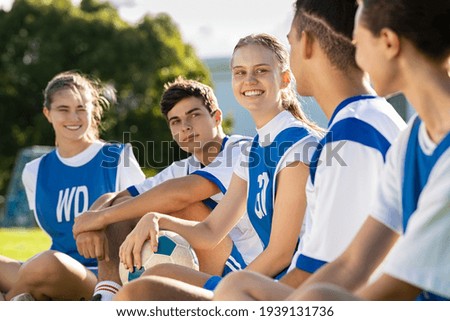 Happy football players sitting on grass on playground field before match. Mixed school soccer players talking during break. Guys and girls teammates sitting in a row for physical education lesson.
