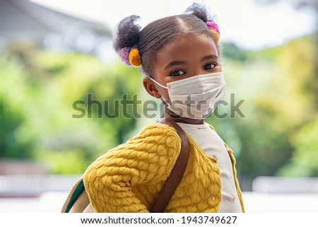 Smiling cute little girl with school backpack and protective face mask ready for first day of school during covid pandemic. Black kid going back to school during coronavirus pandemic disease. 