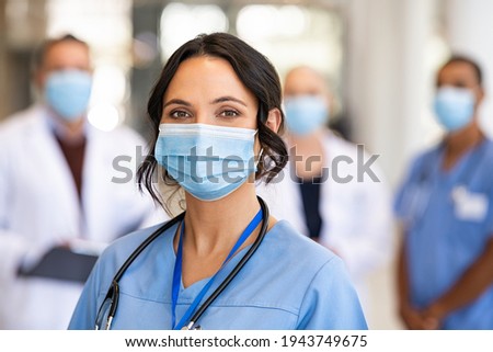 Close up face of confident female nurse in front of his medical staff looking at camera while wearing protective face mask due to covid-19 virus. Smiling surgeon standing  with team in background.