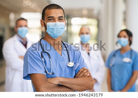 Confident multiethnic male nurse in front of his medical team looking at camera wearing face mask during covid-19 outbreak. Happy and proud indian young surgeon standing in front of his colleagues. 商業照片 © 