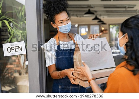 Restaurant owner working only with take away orders during corona virus outbreak. Young black woman wearing face mask giving takeout meal to customer outside her cafeteria. Customer pick up take-away.