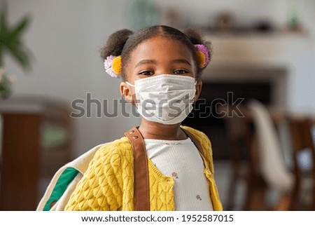Portrait of black little girl wearing bagpack and covid-19 protective face mask ready for elementary school. African female child wearing surgical mask and looking at camera going back to primary.