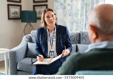 Mature social worker talking to senior man and taking notes of health progress. Mid adult woman in formal clothing visiting old man at home for medical history. Support worker talking to elder.
