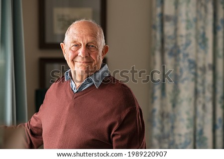 Portrait of happy retired senior man standing at home near window. Satisfied old man looking at camera and smiling while standing near the window. Positive and confident elderly enjoy his retirement.