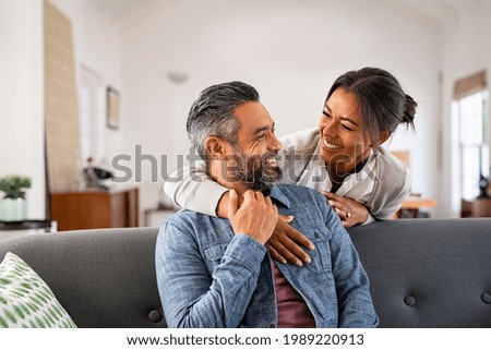 Smiling ethnic woman hugging her husband on the couch from behind in the living room. Middle eastern man having fun with his beautiful young wife on the couch. Mid adult indian man with latin woman.