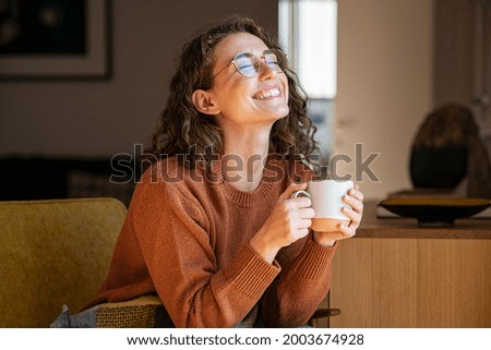Photo of Portrait of joyful young woman enjoying a cup of coffee at home. Smiling pretty girl drinking hot tea in winter. Excited woman wearing spectacles and sweater and laughing in an autumn day.