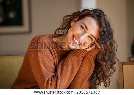Photo of Happy young woman sitting on sofa at home and looking at camera. Portrait of comfortable woman in winter clothes relaxing on armchair. Portrait of beautiful girl smiling and relaxing during autumn.