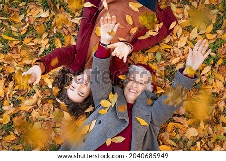 Senior grandmother and smiling granddaughter lying on yellow and red leaves at park, view from above. Top view of playful old woman with cheerful girl lying on autumn park leaves, looking at camera.