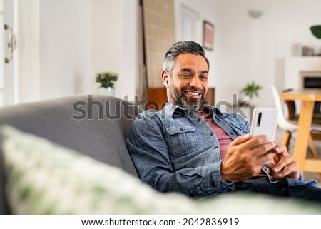 Happy mid adult man using smartphone device while sitting on sofa at home. Smiling mature indian man lying on couch reading messages on mobile phone while listening to music with wireless earbuds. Foto stock © 