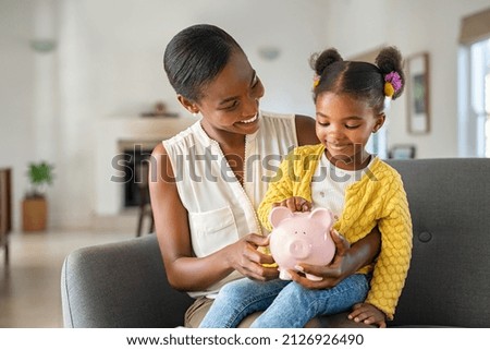Smiling mature african american mother helping daughter sitting on lap putting money in piggy bank. Cute little black girl saving money by adding a coin in piggy bank with mother at home.  Stock foto © 