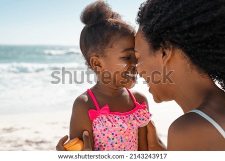 Black mother embracing daughter with sunscreen on face at beach. Woman rubbing nose face to face with her cute little girl with sun lotion on cheek. Funny black female kid with sunblock at seaside. Foto stock © 