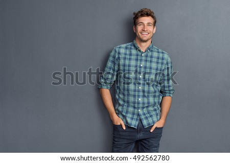 Handsome young man on grey background looking at camera. Portrait of laughing young man with hands in pockets leaning against grey wall. Happy guy smiling. Stock foto © 