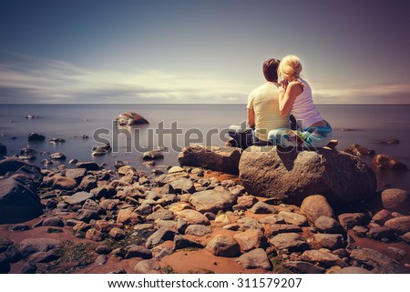 A young couple sitting on a large rock on the seashore and looks at the horizon.