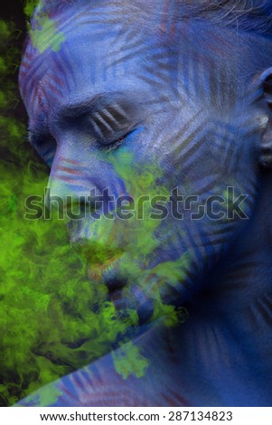 young woman with blue faceart and closed eyes breathing neon toxic green smoke