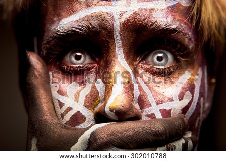 Frightened woman with tribal face painting and blue eyes art holding her mouth with a hand and not allowing herself to scream or talk