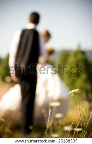 An out of focus bride and groom walk through a field with flowers and trees after their wedding.