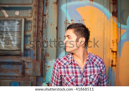 A mid-20s man is outside of a train car with graffiti modeling a plaid western button up shirt.