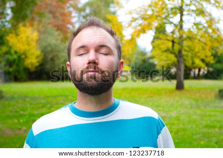 A man with a full beard closes his eyes while the camera photographs him in a park setting outdoors.