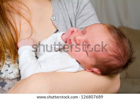 A mother holds her newborn baby in her arms while looking down at him with a happy look. The baby boy is in comfort and trusts his mom fully. He is sleeping peaceful.y.