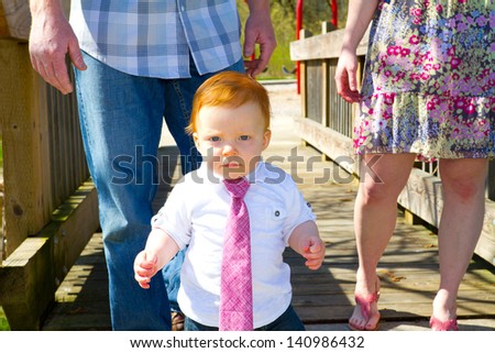 A baby wearing a red or pink necktie walks across a bridge with his parents behind him.