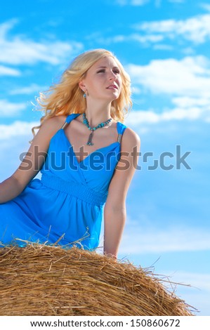 Beautiful young woman in blue dress  is sitting on stack of hay on sunny day in the country
