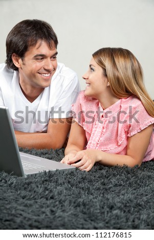 Happy young couple looking at each other while lying on rug with laptop