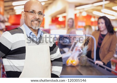Portrait of a grocery store cashier standing at checkout counter with customers in the background