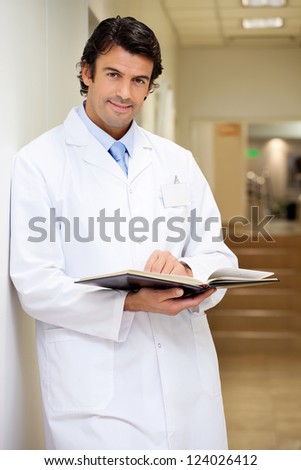 Portrait of a mixed race male doctor holding book while standing in hospital corridor