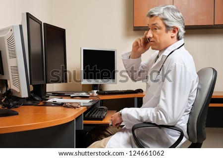 Mature male doctor attending phone call while sitting in front of computer desk at clinic