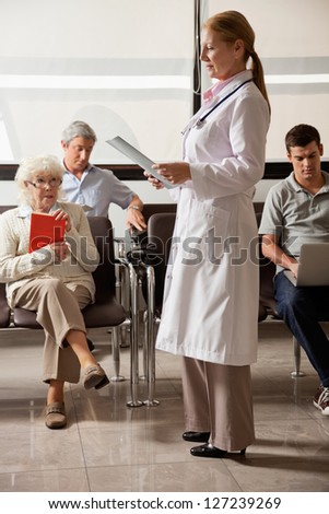 Full length of female doctor reading file with people sitting in hospital lobby