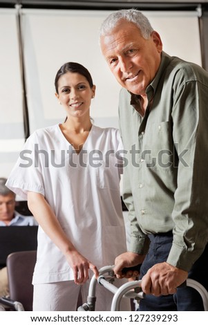 Portrait of nurse helping senior male patient to use walker with person sitting in background