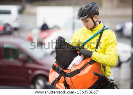 Young male cyclist in protective gear putting package in courier bag on street