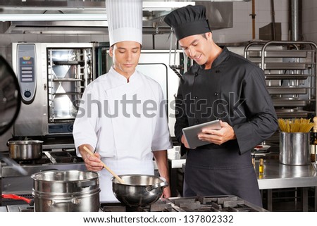 Male chef with digital computer assisting colleague in preparing food at kitchen