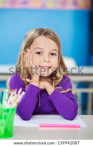 Portrait of cute little girl sitting with hand on chin at desk in kindergarten