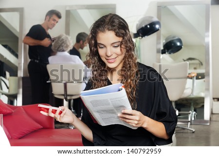 Young woman holding mobile phone while reading magazine with hairdresser and senior woman in the background at hair salon