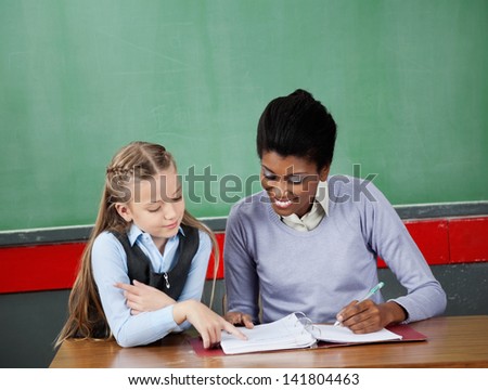 Little schoolgirl asking question to female teacher at desk in classroom