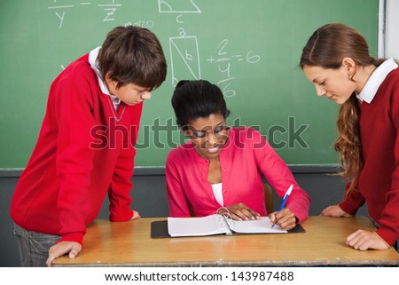Young female teacher writing in binder while students standing at desk in classroom