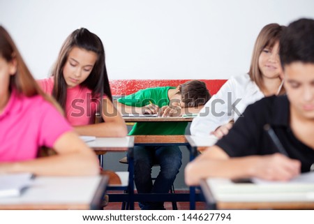 Teenage male student sleeping on desk with friends in foreground at classroom