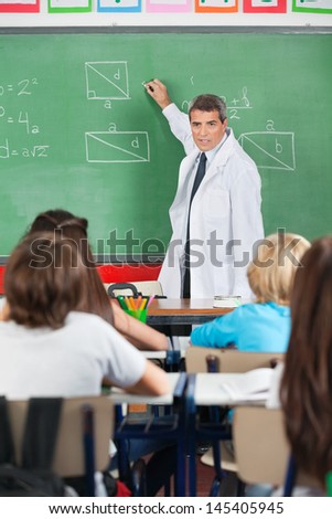 Mature male teacher teaching while writing on board with students in foreground at classroom