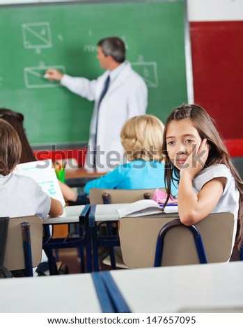 Bored little schoolgirl sitting at desk with teacher teaching geometry in classroom